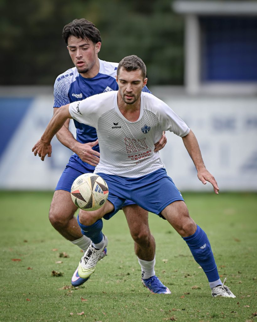 Fußballspieler in Weiß schirmt den Ball gegen einen Spieler in Blau ab