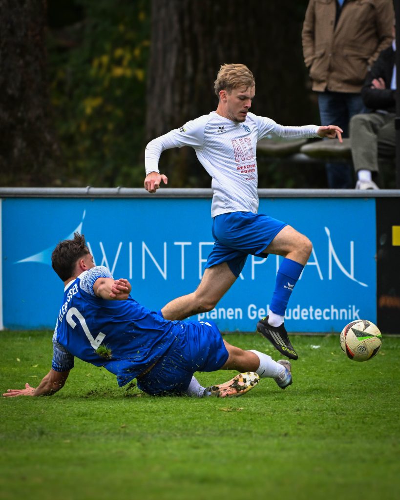 Spieler in weißem Trikot springt über einen grätschenden Gegenspieler in Blau
