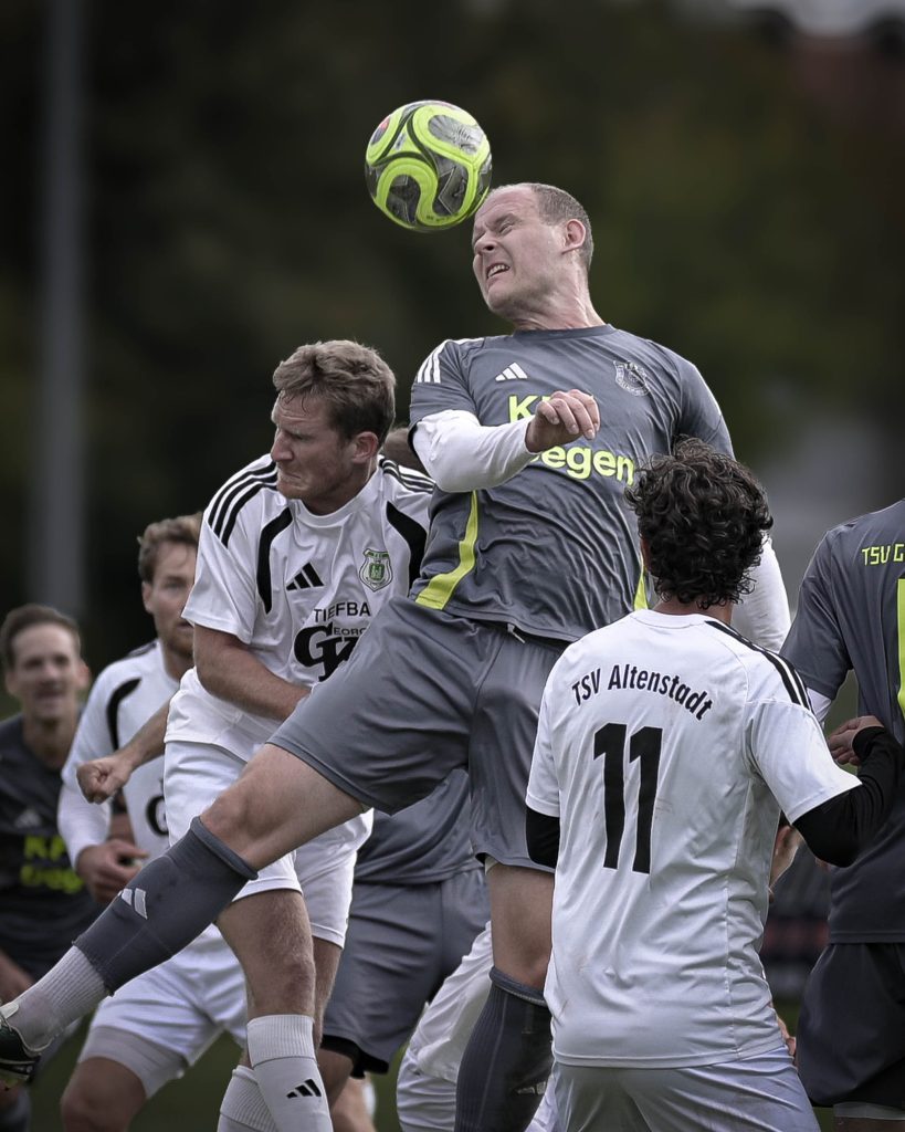 Fußballspieler in grauem Trikot steigt zwischen weißen Gegenspielern zum Kopfball