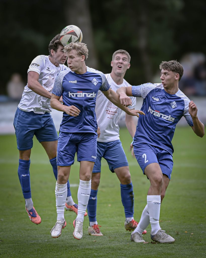 Vier Fußballspieler in weißen und blauen Trikots verfolgen einen hohen Ball