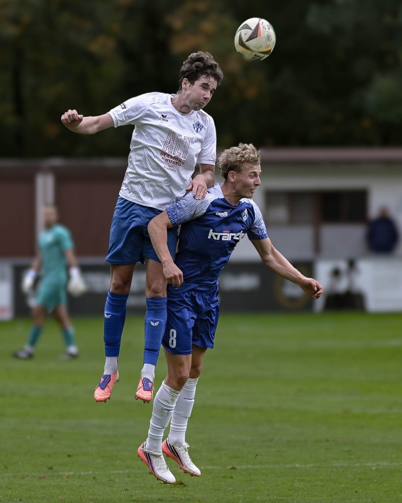 Spieler in weißem Trikot steigt hinter einem blauen Gegenspieler zum Kopfball hoch
