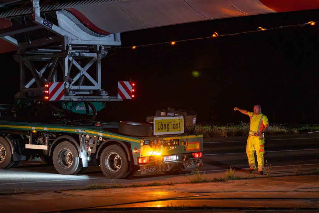 Einweiser in Warnkleidung gibt neben dem Auflieger eines Rotorblatttransports ein Handzeichen