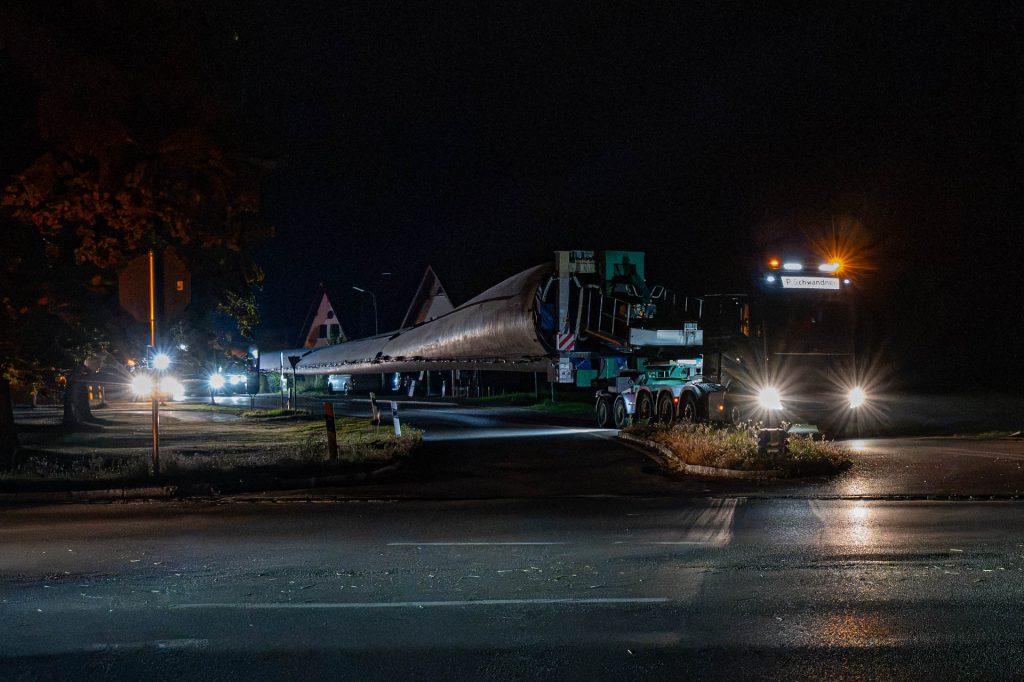 Zugmaschine mit langem Rotorblatt fährt nachts an einer Verkehrsinsel und Häusern vorbei
